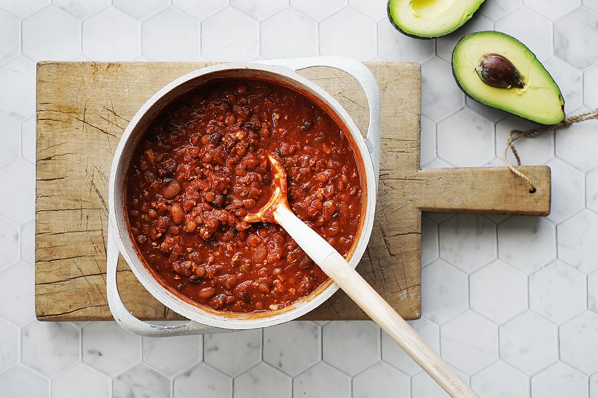 A saucepan with beans and tofu chili and an avocado on the side.