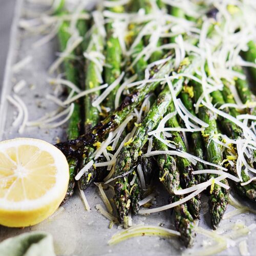 Grilled asparagus, with parmesan cheese on top, and lemon on the side, under a metal base.