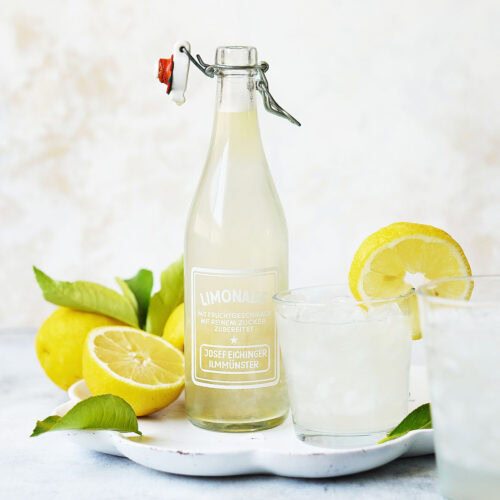 Lemonade in a glass container, next to a glass full of lemonade, on the side fresh lemons.