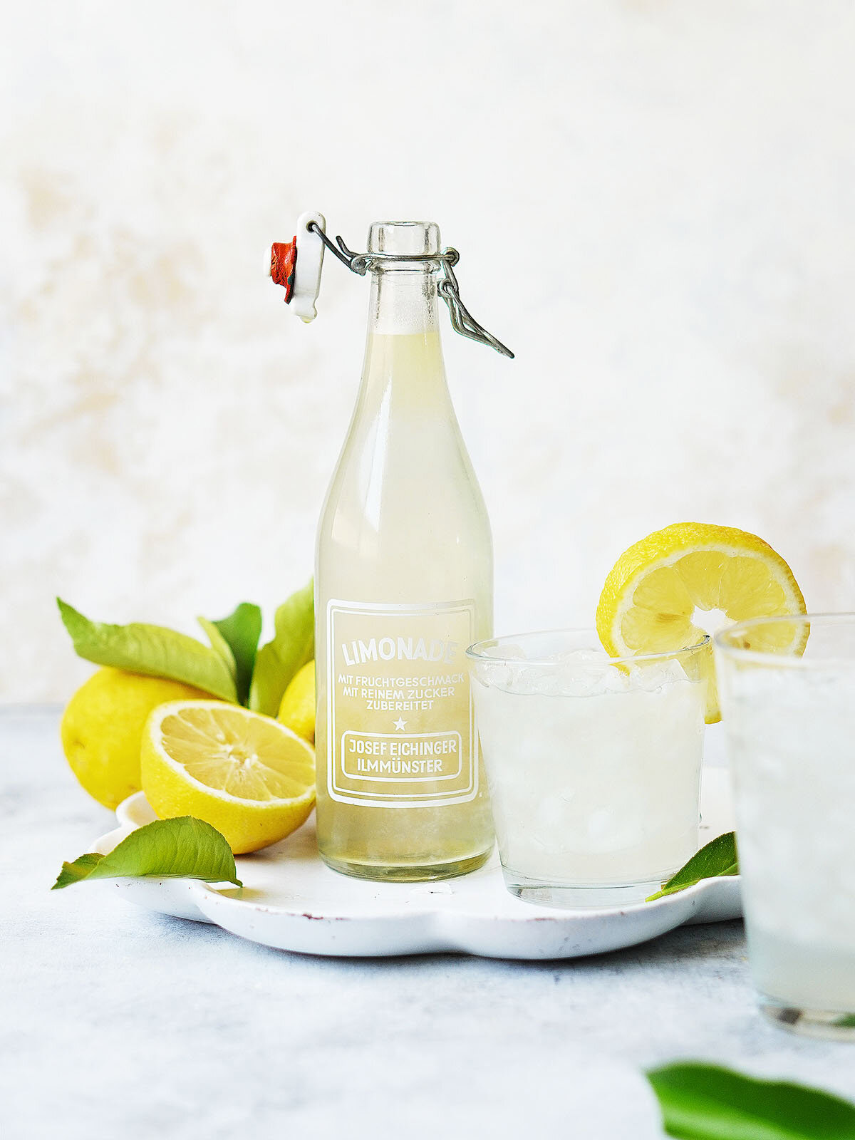 Lemonade in a glass container, next to a glass full of lemonade, on the side fresh lemons.
