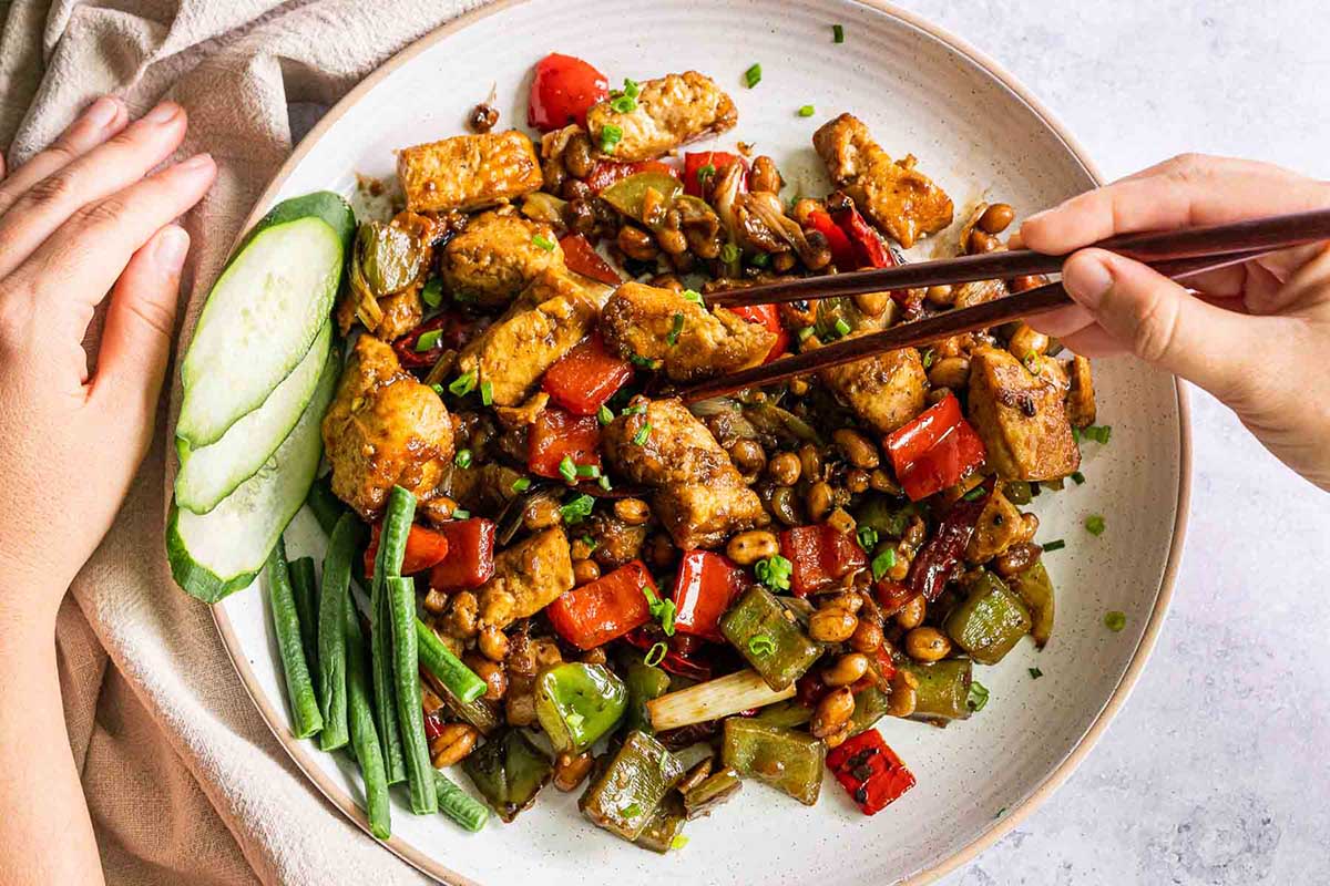 Kung pao pork on a plate with hands holding chopsticks.