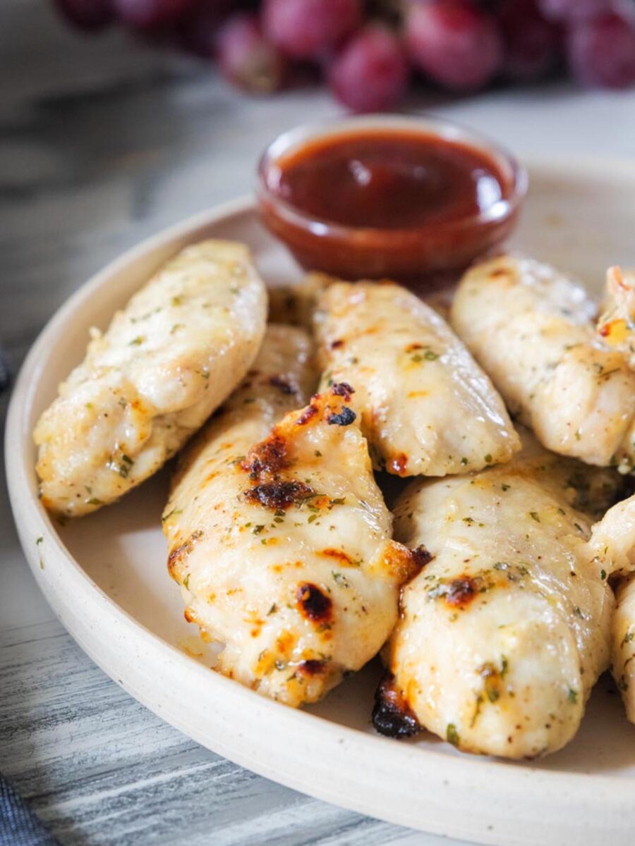 Plate of grilled chicken tenders with herbs, served with a small dish of red dipping sauce. Grapes are blurred in the background.