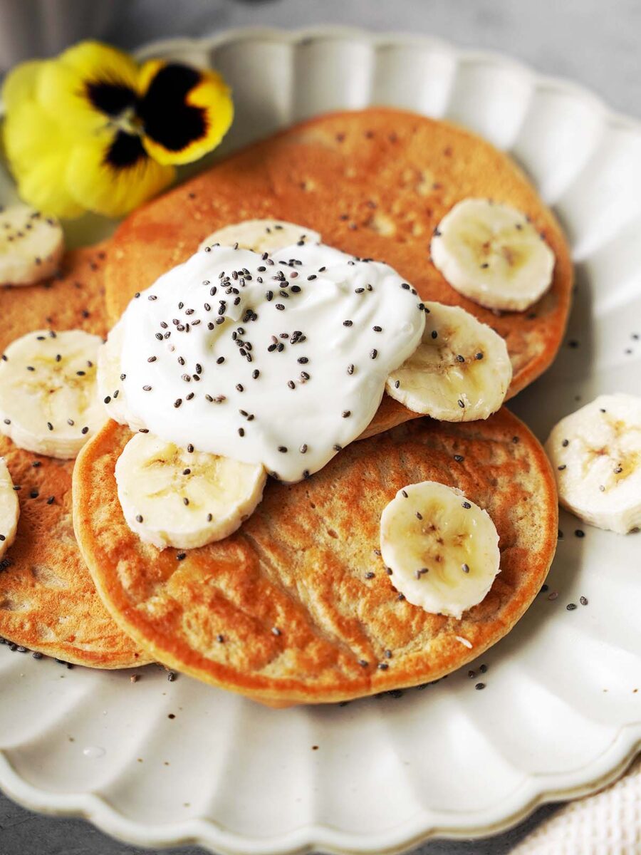 Three pancakes on a white plate, topped with banana slices, a dollop of yogurt, chia seeds, and garnished with a yellow pansy flower.