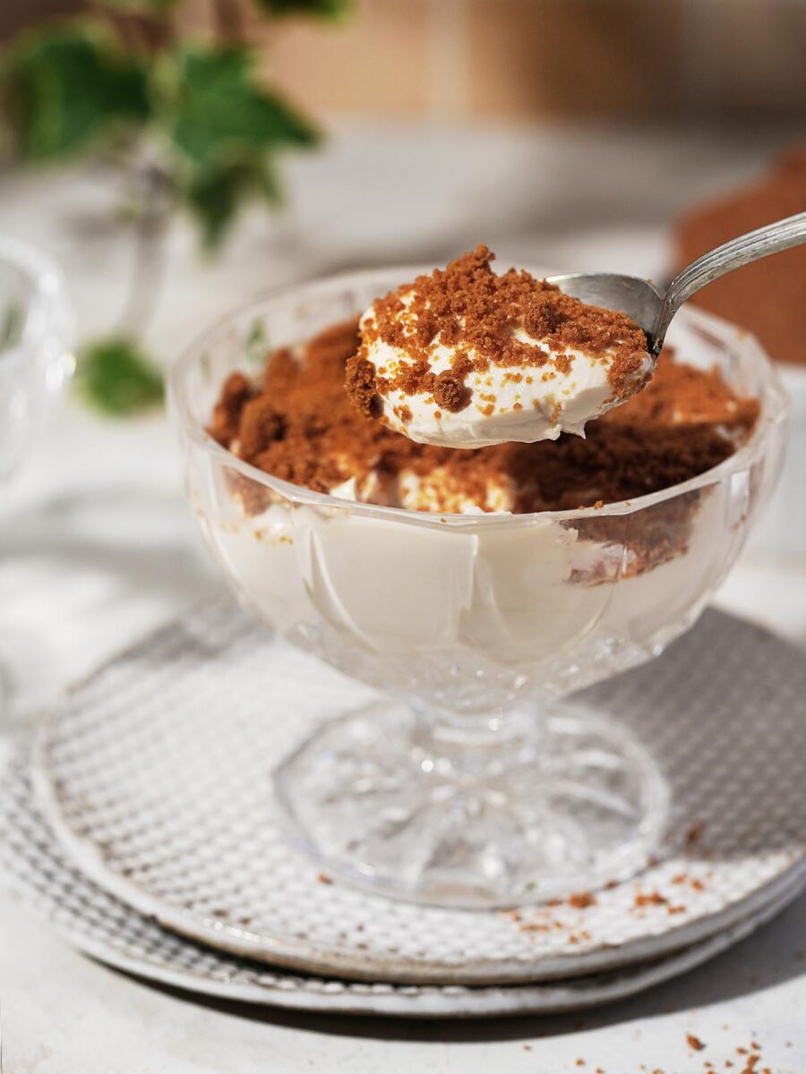 A glass dessert dish filled with creamy Protein Cheesecake topped with crumbled brown cookies, with a spoon lifting a bite of the dessert above the dish.