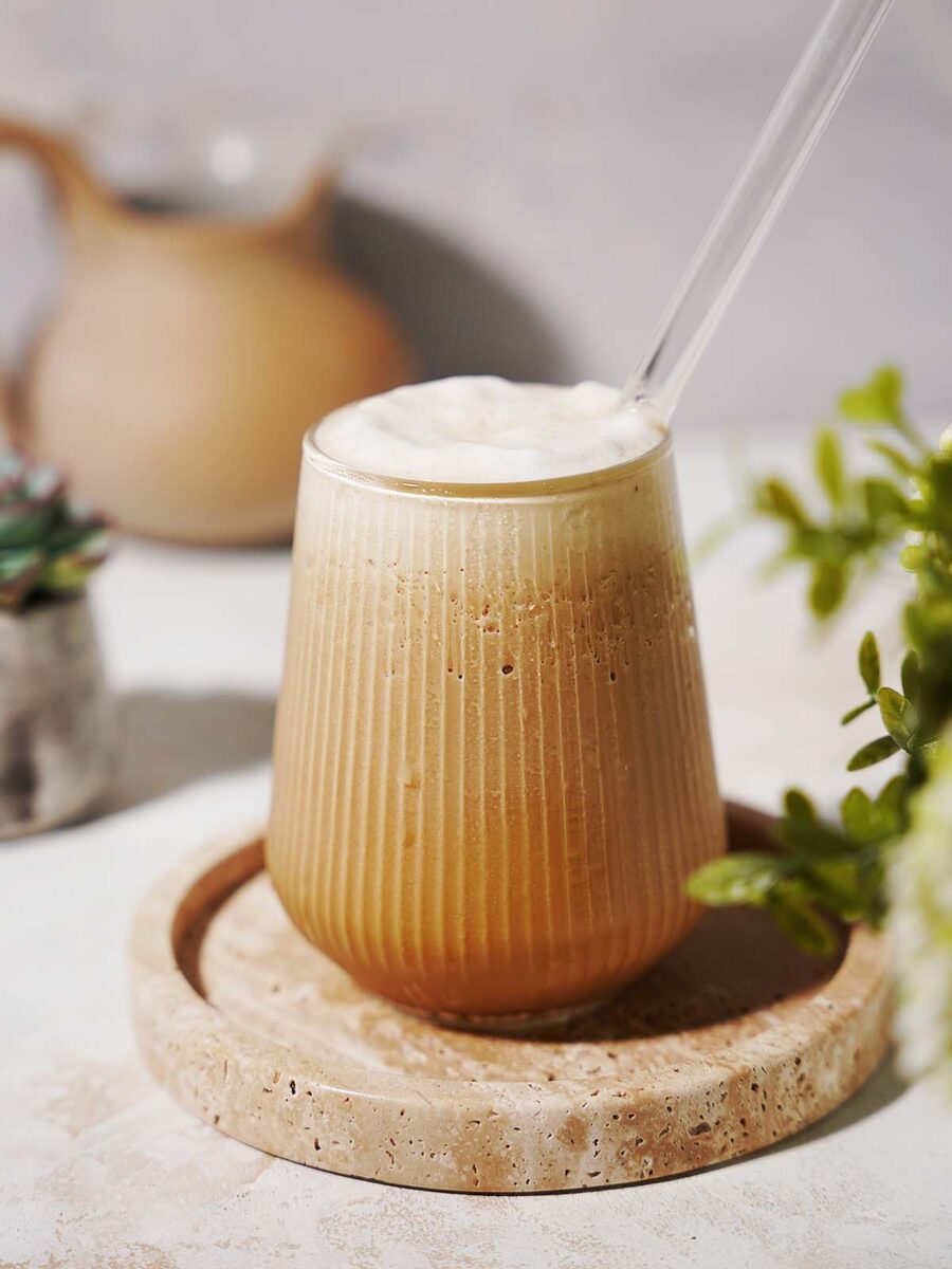 A glass of iced coffee with frothy foam on top, served with a clear straw on a round stone coaster. Green plants and a blurred jug are in the background.