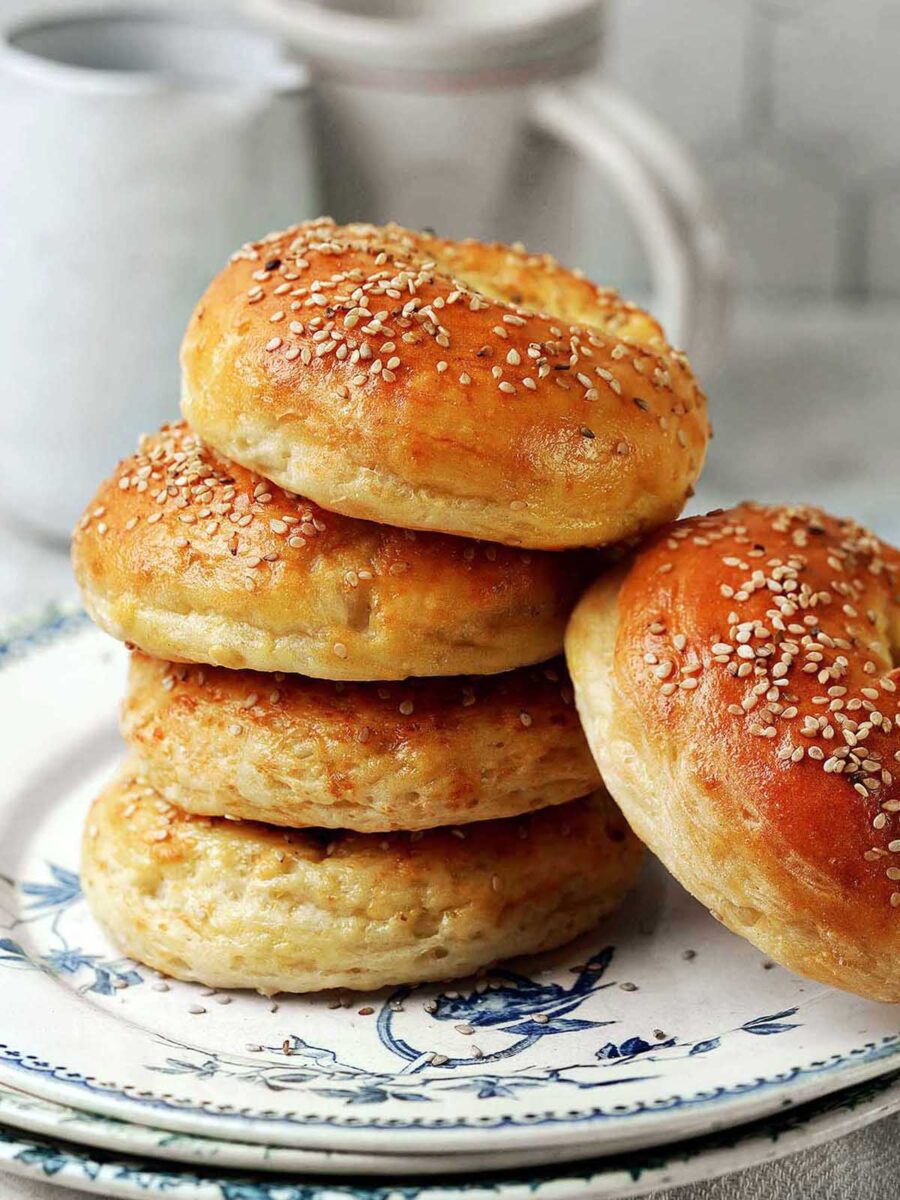 A stack of golden, sesame seed-topped protein bagels sits on a decorative plate, with a blurred white mug and pitcher in the background.