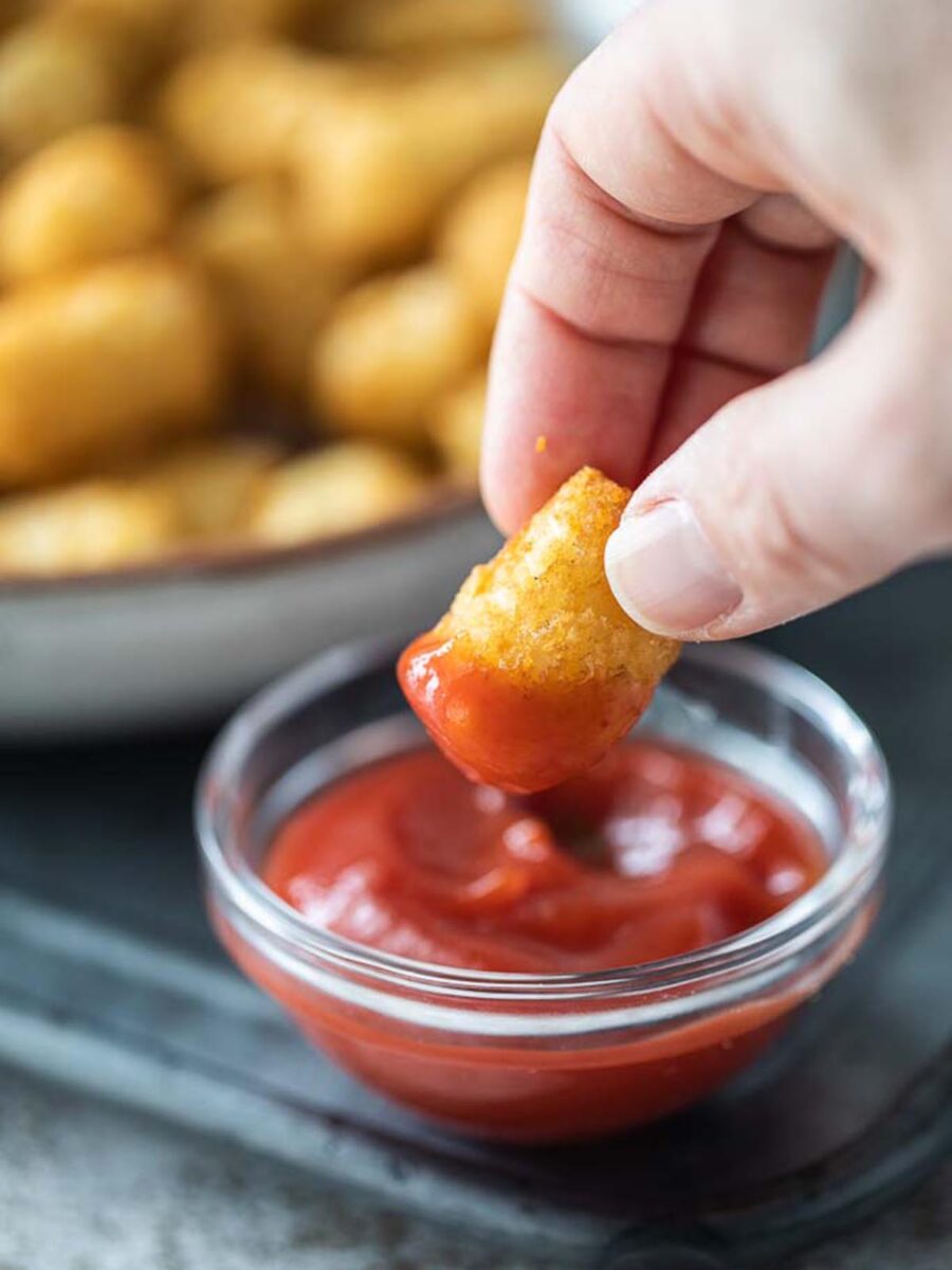 A hand with a tater tot dipping it into a small bowl with ketchup.