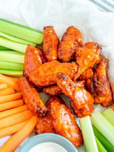 A pile of orange buffalo chicken wings served with carrot sticks, celery stalks, and a small bowl of dipping sauce.