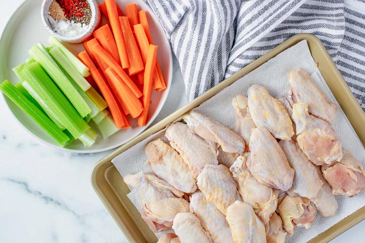 Raw chicken wings arranged on a parchment-lined baking sheet next to a plate of celery and carrot sticks with a small dish of seasoning.