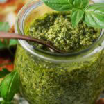 Fresh homemade green pesto in glass jar with spoonful of pesto, basil leaves, cheese, and walnuts on a patterned tray, healthy and delicious sauce.