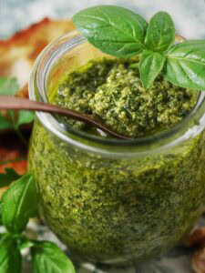 Fresh homemade green pesto in glass jar with spoonful of pesto, basil leaves, cheese, and walnuts on a patterned tray, healthy and delicious sauce.