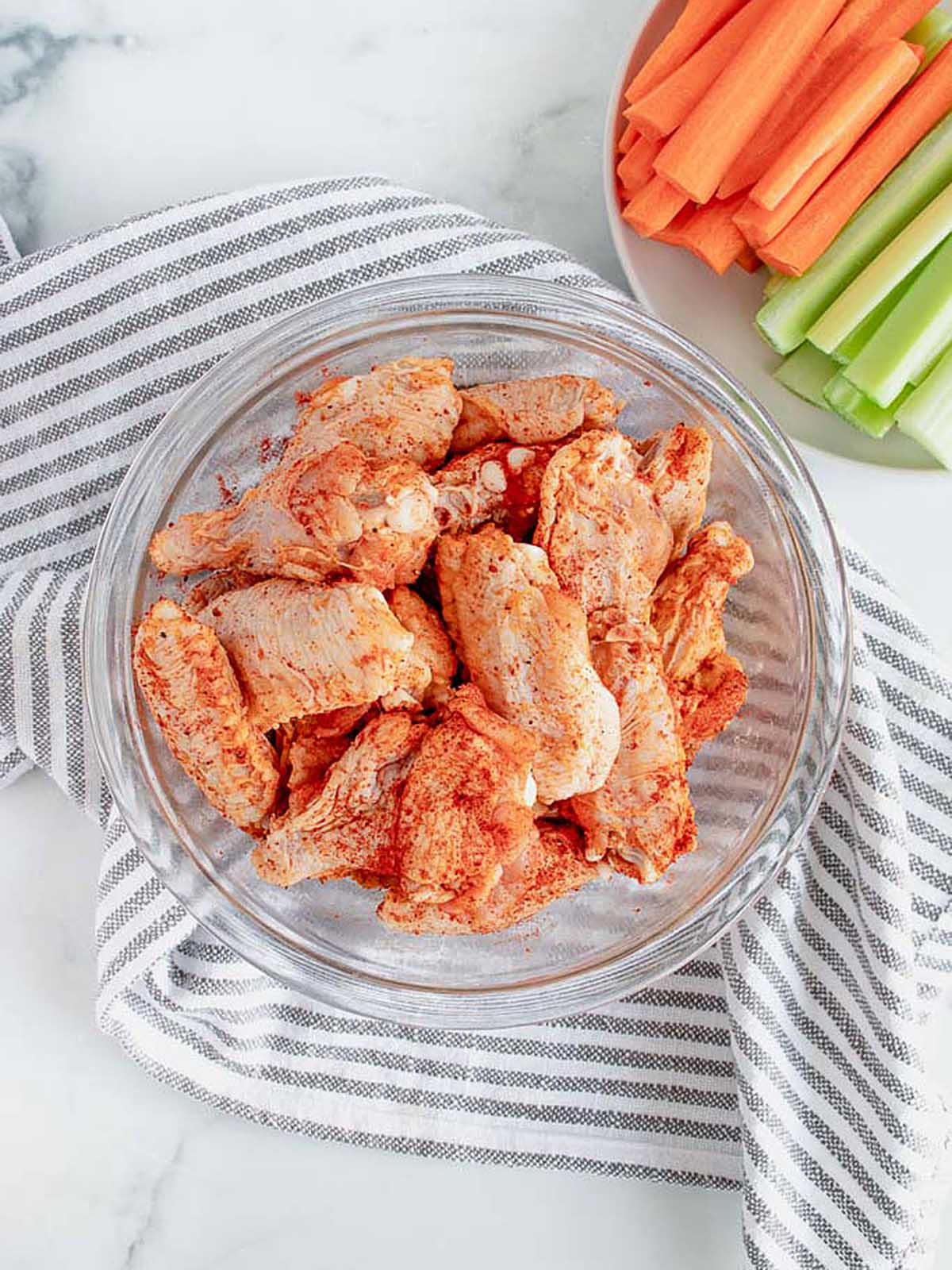 A glass bowl filled with raw chicken wings seasoned with paprika, next to a plate of carrot and celery sticks.