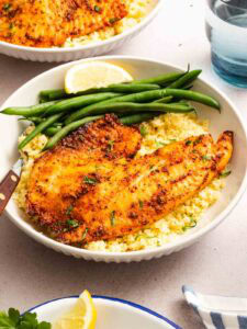 A bowl with a seasoned, cooked fish fillet on a bed of couscous, accompanied by green beans and a lemon wedge. A knife rests on the side, and a glass of water is visible in the background.