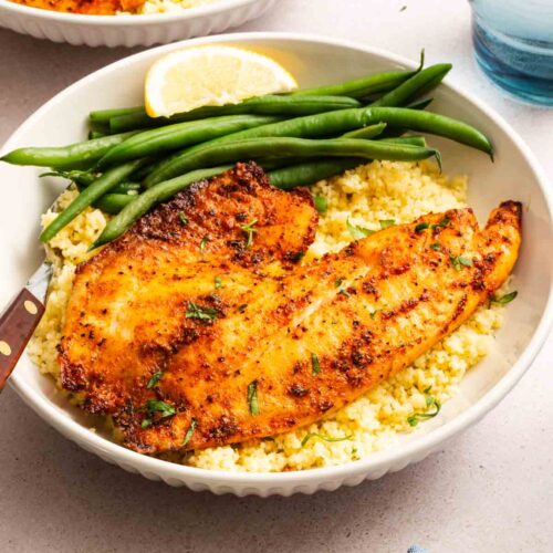 A bowl with a seasoned, cooked fish fillet on a bed of couscous, accompanied by green beans and a lemon wedge. A knife rests on the side, and a glass of water is visible in the background.