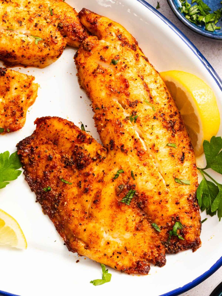 Seasoned, pan-seared fish fillets garnished with fresh herbs on a white plate, served with lemon wedges and parsley.