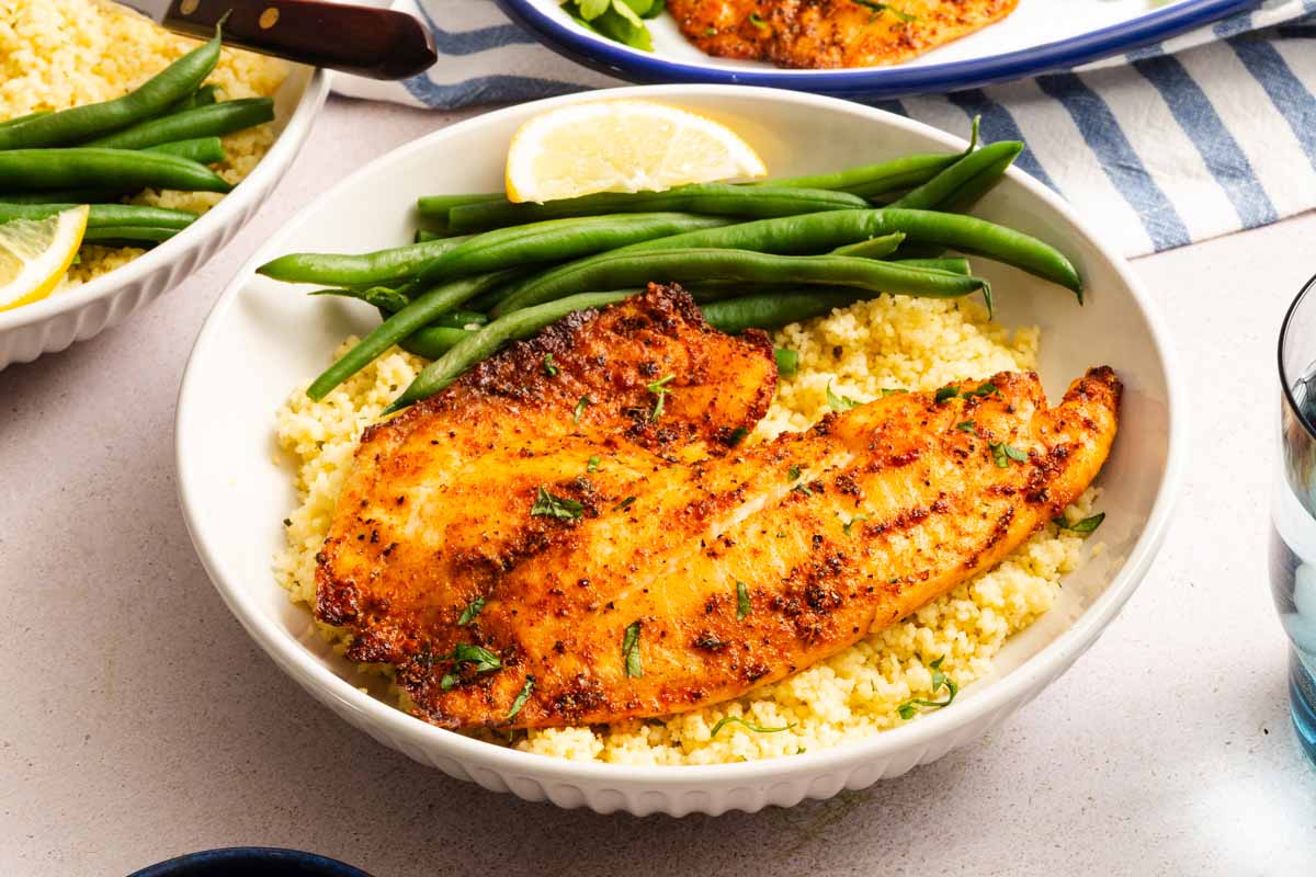 A bowl with seasoned, cooked fish fillets served over couscous, accompanied by green beans and a lemon wedge.
