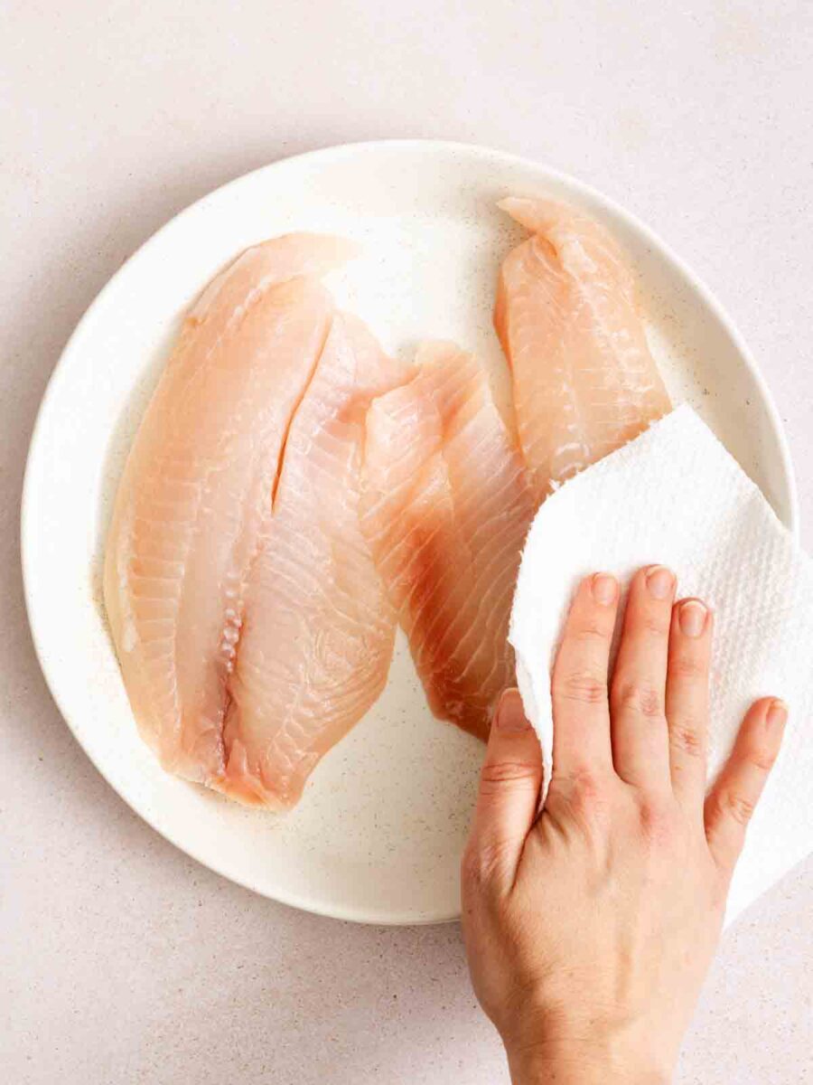 A hand uses a paper towel to pat dry raw fish fillets on a white plate.