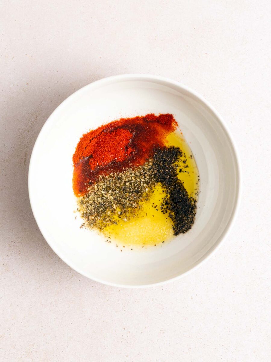 A white bowl containing separate piles of yellow oil, black pepper, dried herbs, red paprika, and white granulated garlic or salt, arranged on a light surface.