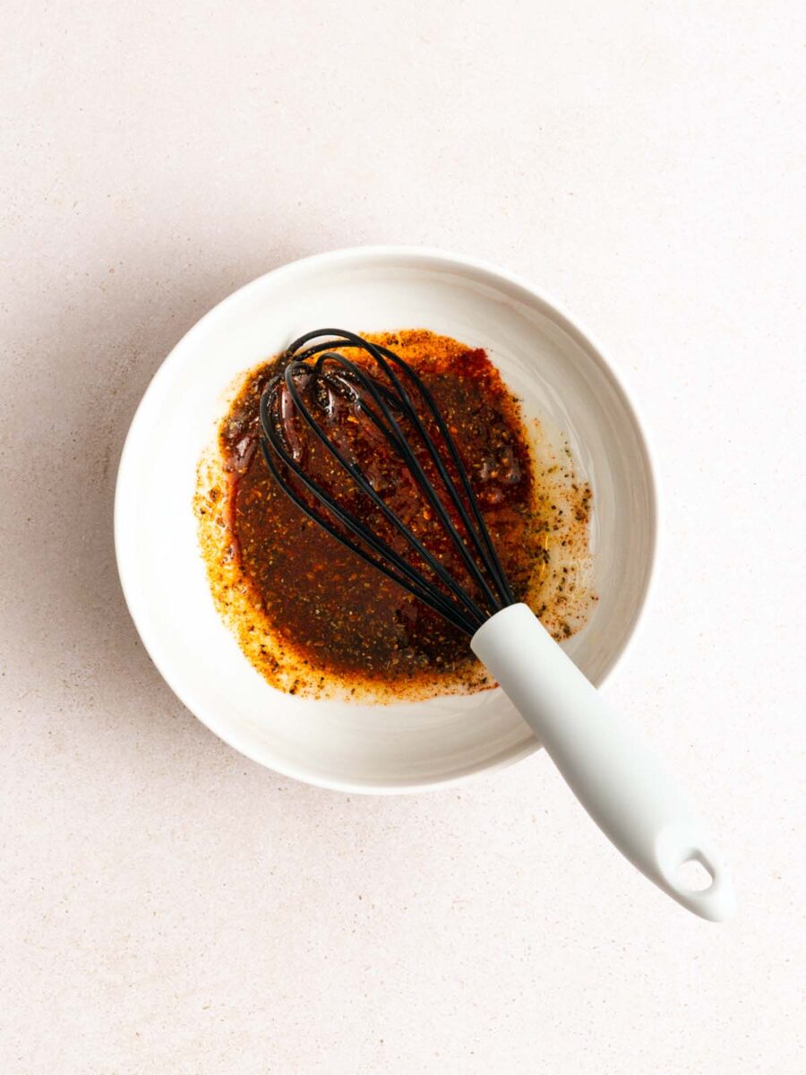 A white bowl containing a brown, seasoned liquid mixture being whisked with a white-handled whisk on a light-colored surface.