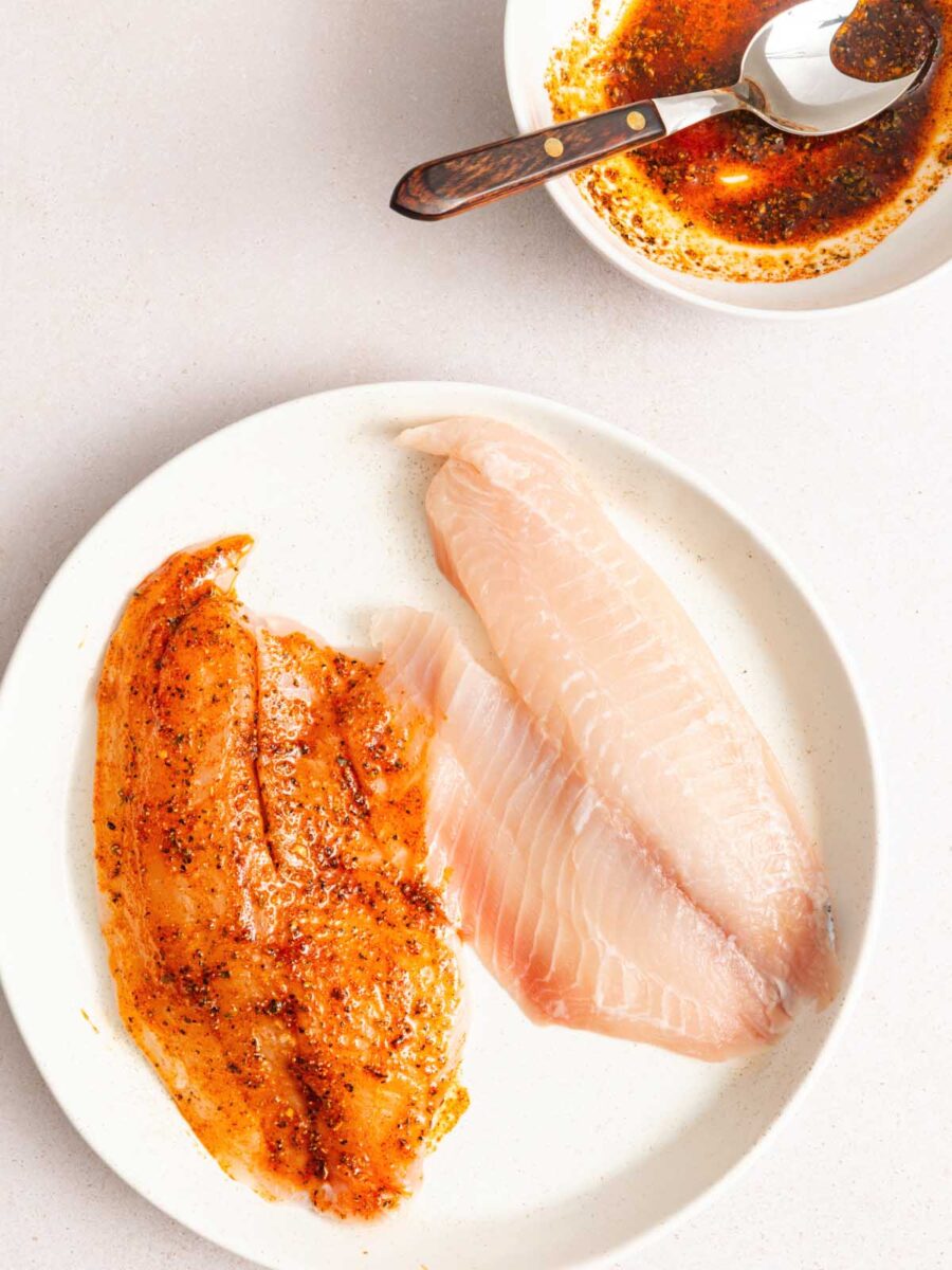 Two raw fish fillets on a white plate; one is coated with a reddish-brown seasoning while the other is plain. Above, a bowl contains more seasoning mixture with a spoon resting in it.