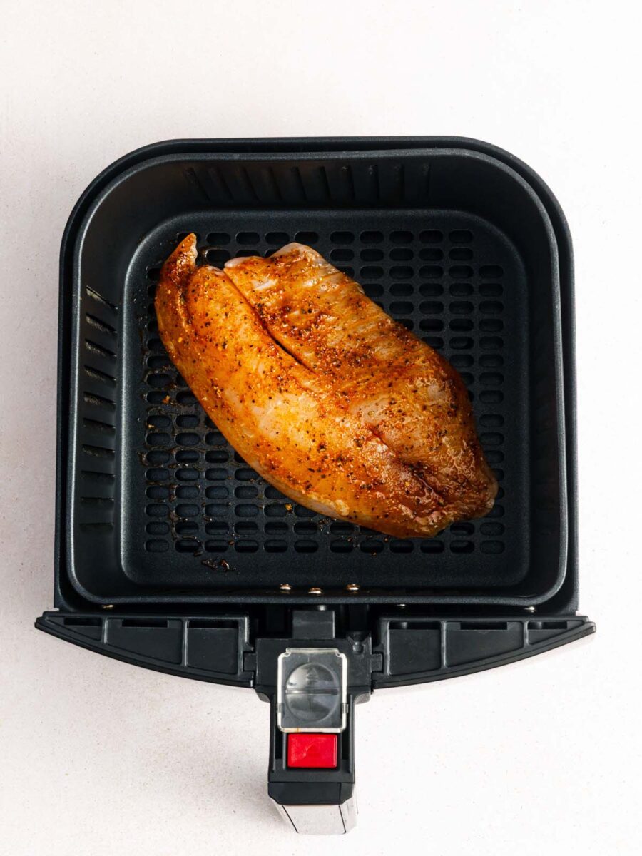 A cooked, seasoned chicken breast sits in the basket of an open air fryer on a light-colored surface.
