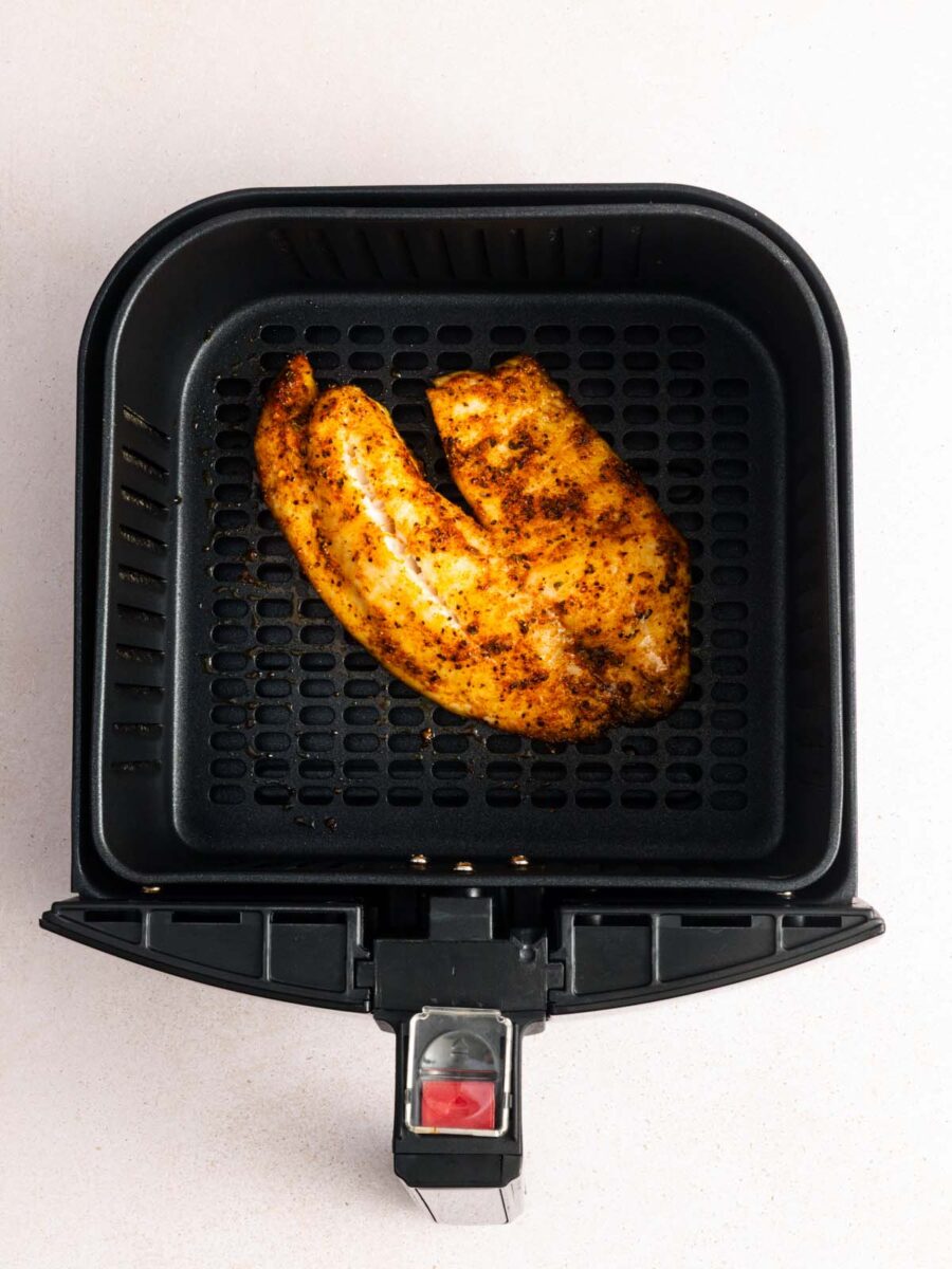A seasoned chicken breast fillet cooks inside a black air fryer basket, viewed from above on a light-colored surface.