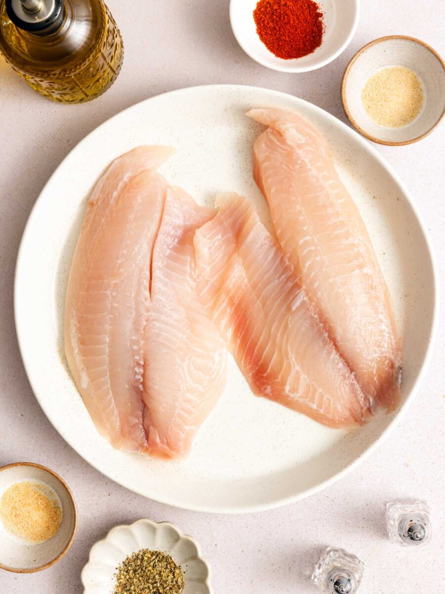 Two raw fish fillets are placed on a white plate, surrounded by small bowls of spices, a bottle of olive oil, and salt and pepper shakers on a light-colored surface.