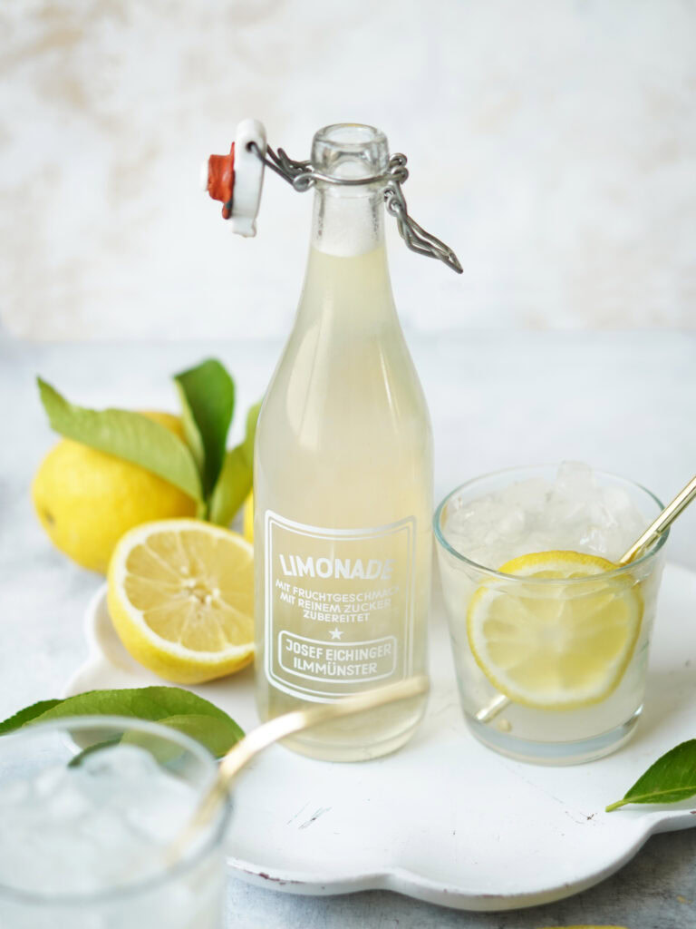 A glass bottle of lemonade with a swing-top lid stands on a white tray, flanked by two glasses of lemonade with ice and lemon slices. Fresh lemons and green leaves are placed beside the bottle.