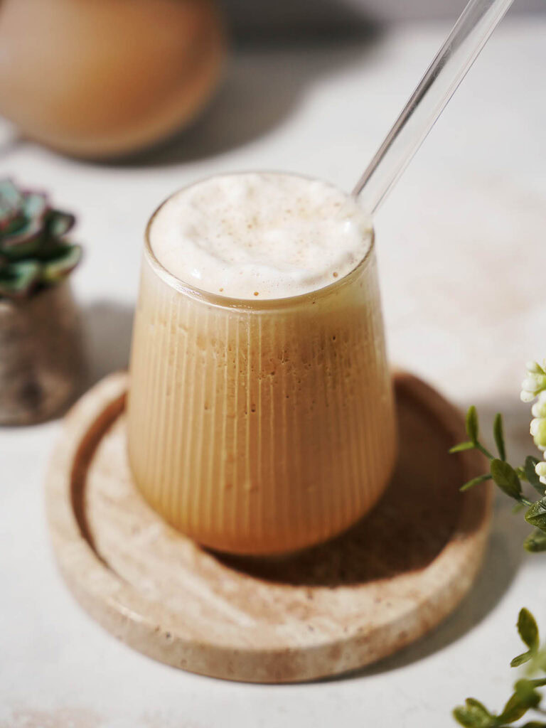A glass of iced coffee with frothy foam on top and a clear straw, placed on a round stone coaster. A small succulent plant and white flowers are visible nearby on a light surface.