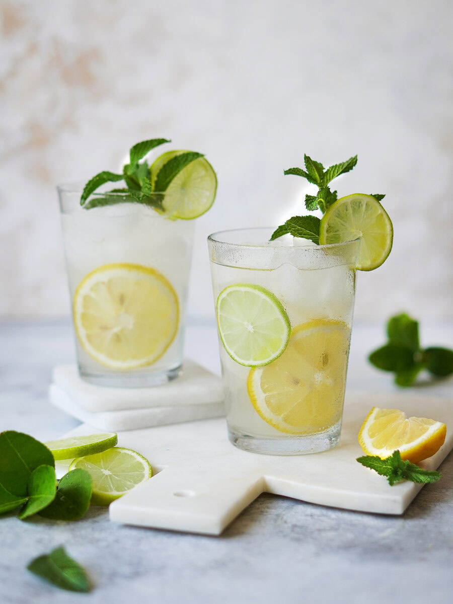 Two glasses of lemonade with ice, lemon and lime slices, and mint sprigs sit on white marble boards. Lemon wedges, lime slices, and mint leaves are scattered around on a light surface.