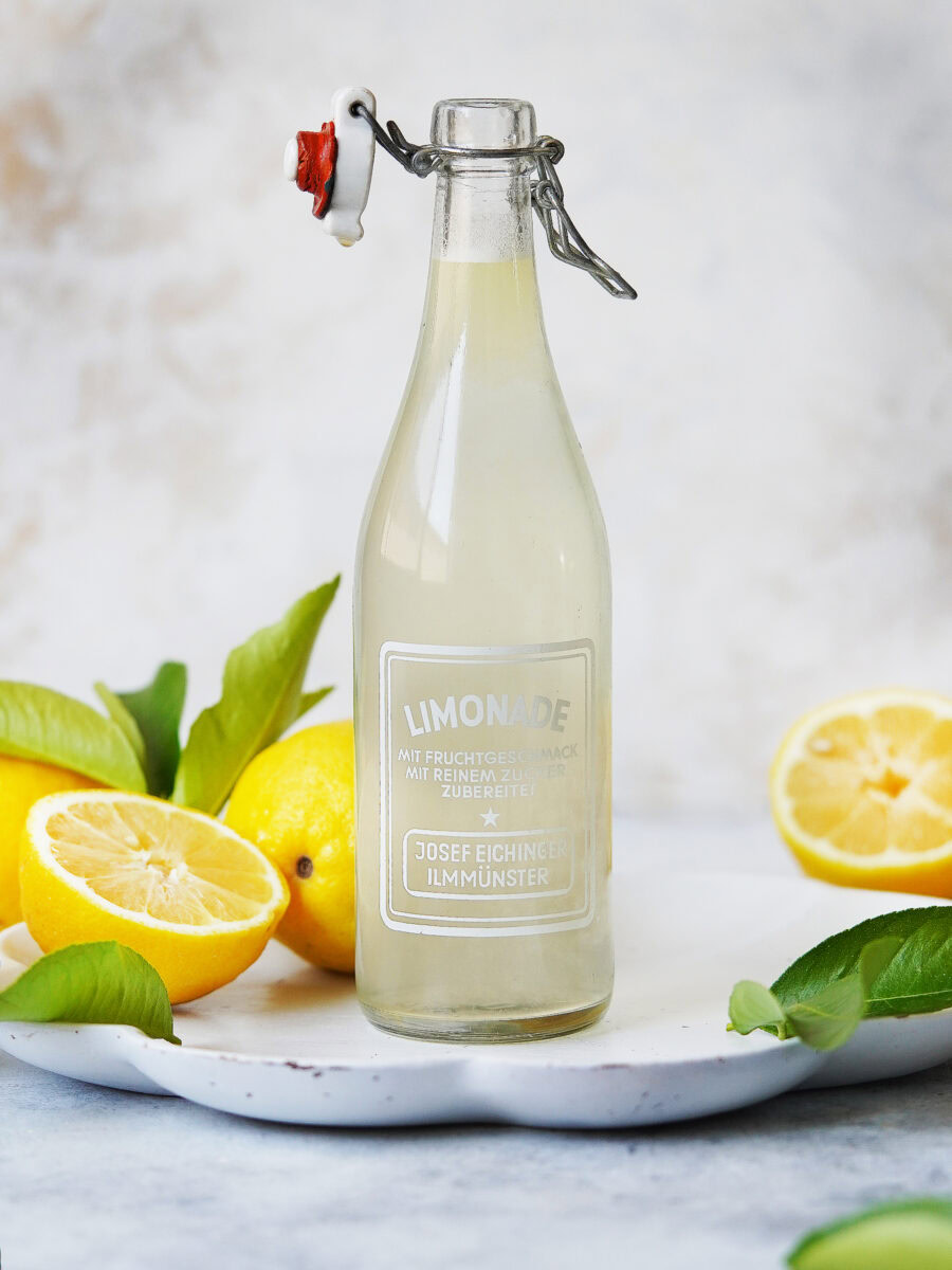 A glass bottle of lemonade with a swing top, surrounded by whole and halved lemons and green leaves, sits on a white plate against a light, blurred background.