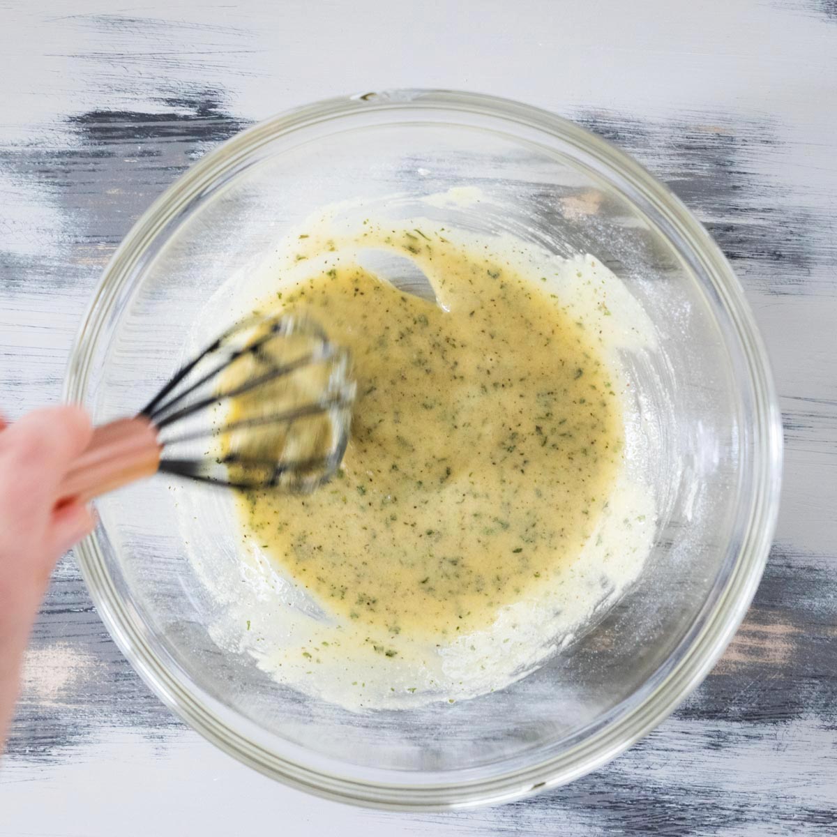 A hand whisks a yellowish mixture with herbs in a clear glass bowl on a light gray surface.