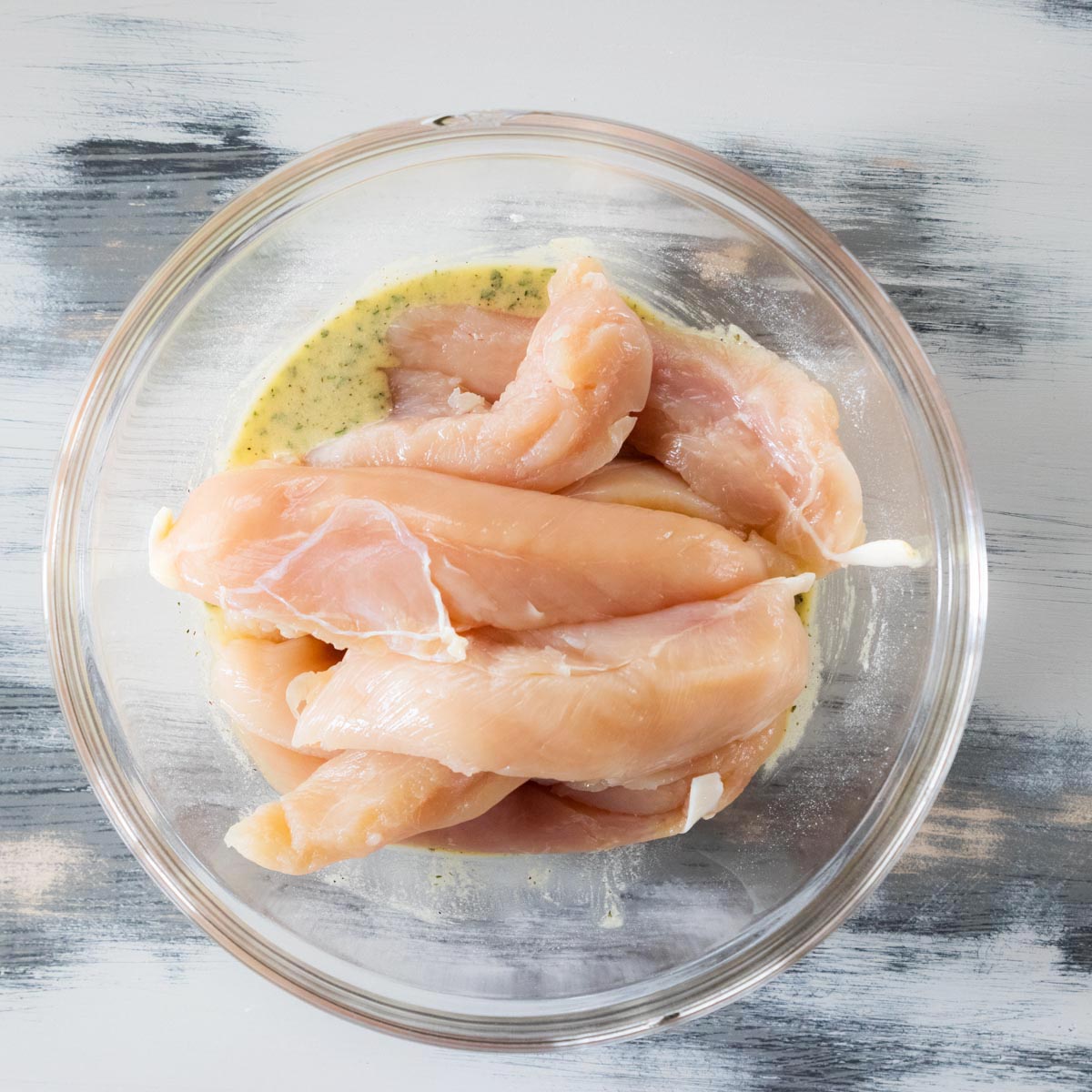 Raw chicken tenders marinating in a clear glass bowl with a light-colored seasoning mixture, placed on a gray and white wooden surface.