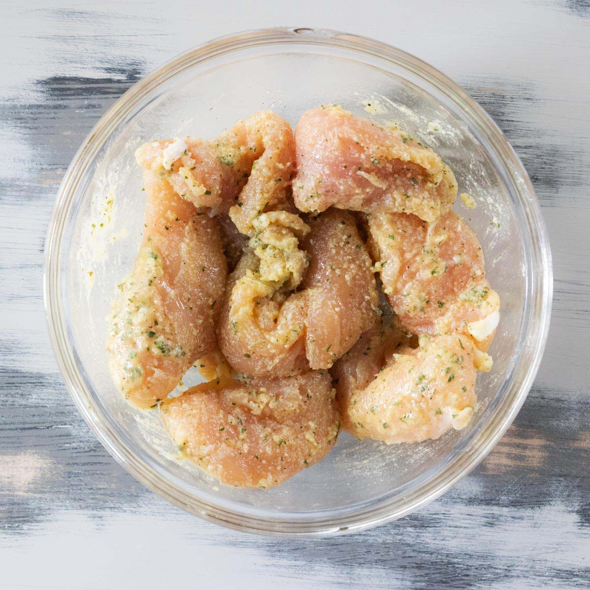 Raw chicken strips coated in a seasoned marinade are arranged in a clear glass bowl, sitting on a light gray wooden surface with visible brush strokes.