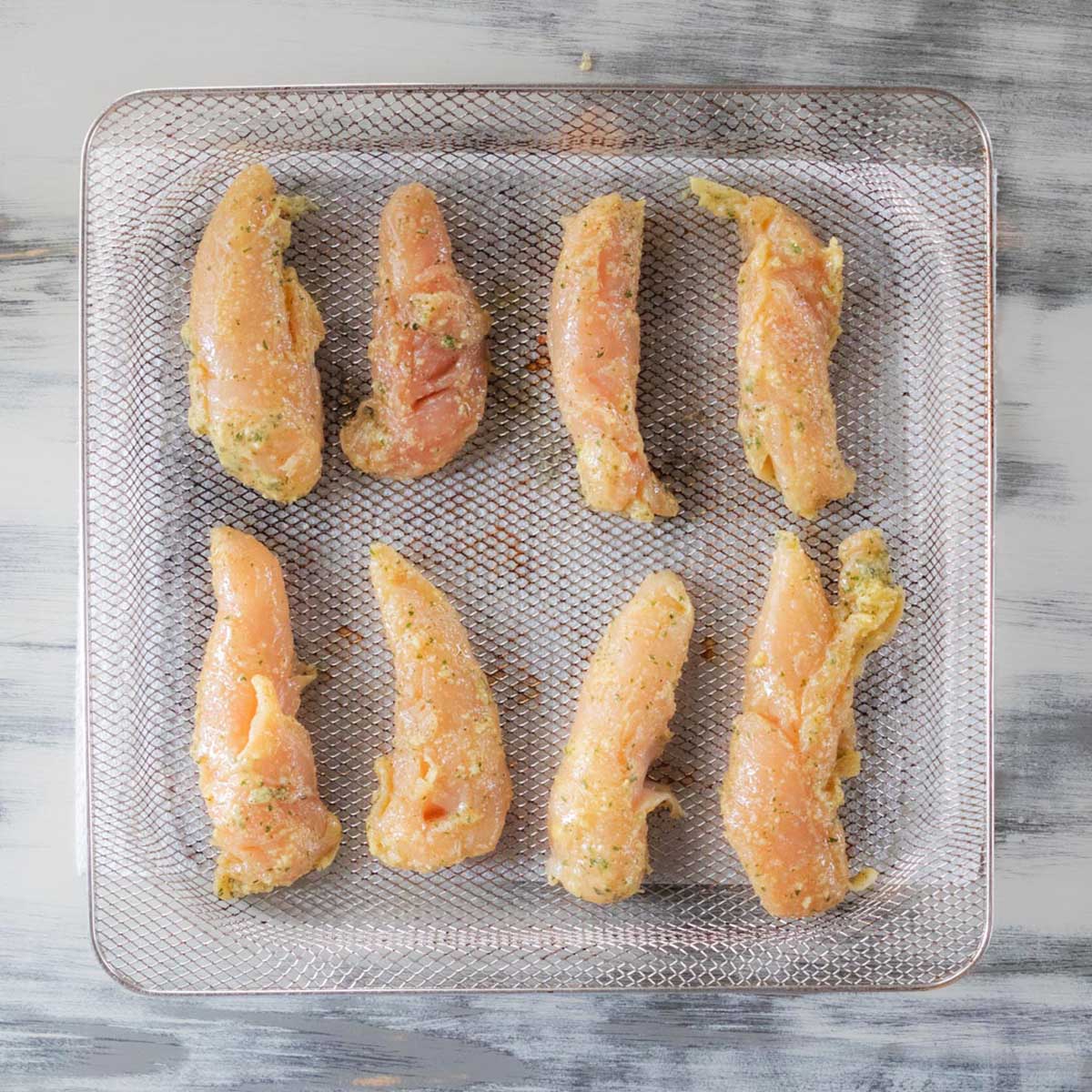 Eight raw, seasoned chicken tenders are evenly spaced in a single layer on a metal air fryer basket, ready to be cooked. The background is a light, rustic wooden surface.