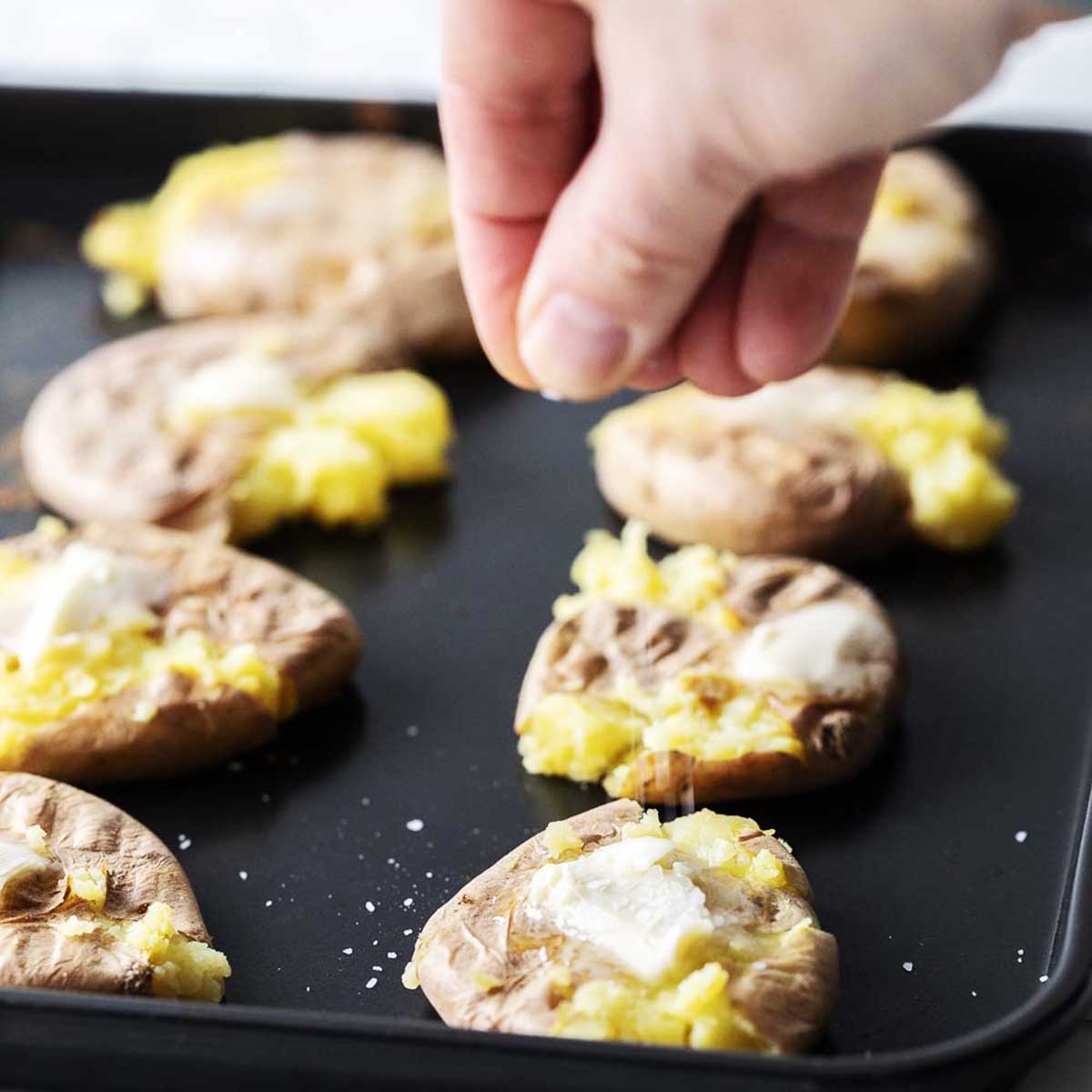 A hand sprinkles salt over smashed potatoes with pats of butter on a black baking tray.