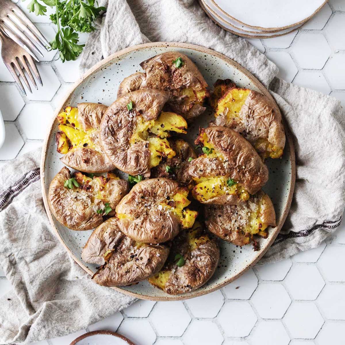 A plate of crispy smashed potatoes garnished with herbs sits on a neutral cloth atop a white hexagonal tiled surface, with forks and parsley nearby.