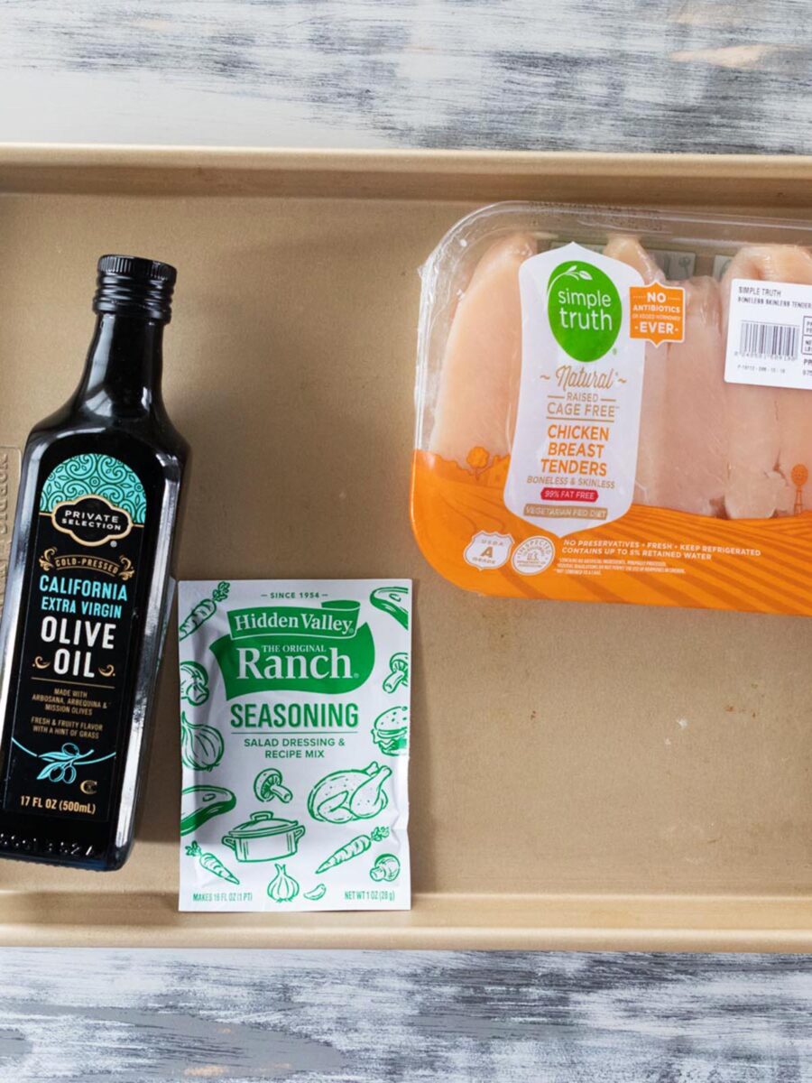 A tray with a bottle of olive oil, a packet of ranch seasoning mix, and a package of raw chicken breast tenders is placed on a wood-textured surface.