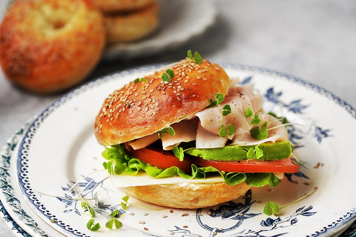 A protein bagel sandwich with lettuce, tomato, avocado, turkey slices, and microgreens sits on a decorative plate, with more bagels blurred in the background.