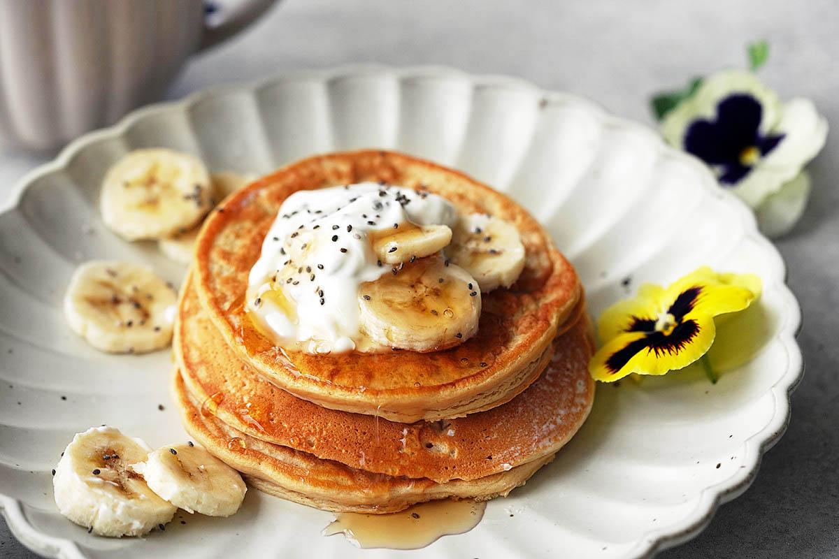 A stack of pancakes topped with banana slices, yogurt, chia seeds, and syrup on a white plate, garnished with more banana slices and a yellow pansy flower. A mug is partially visible in the background.