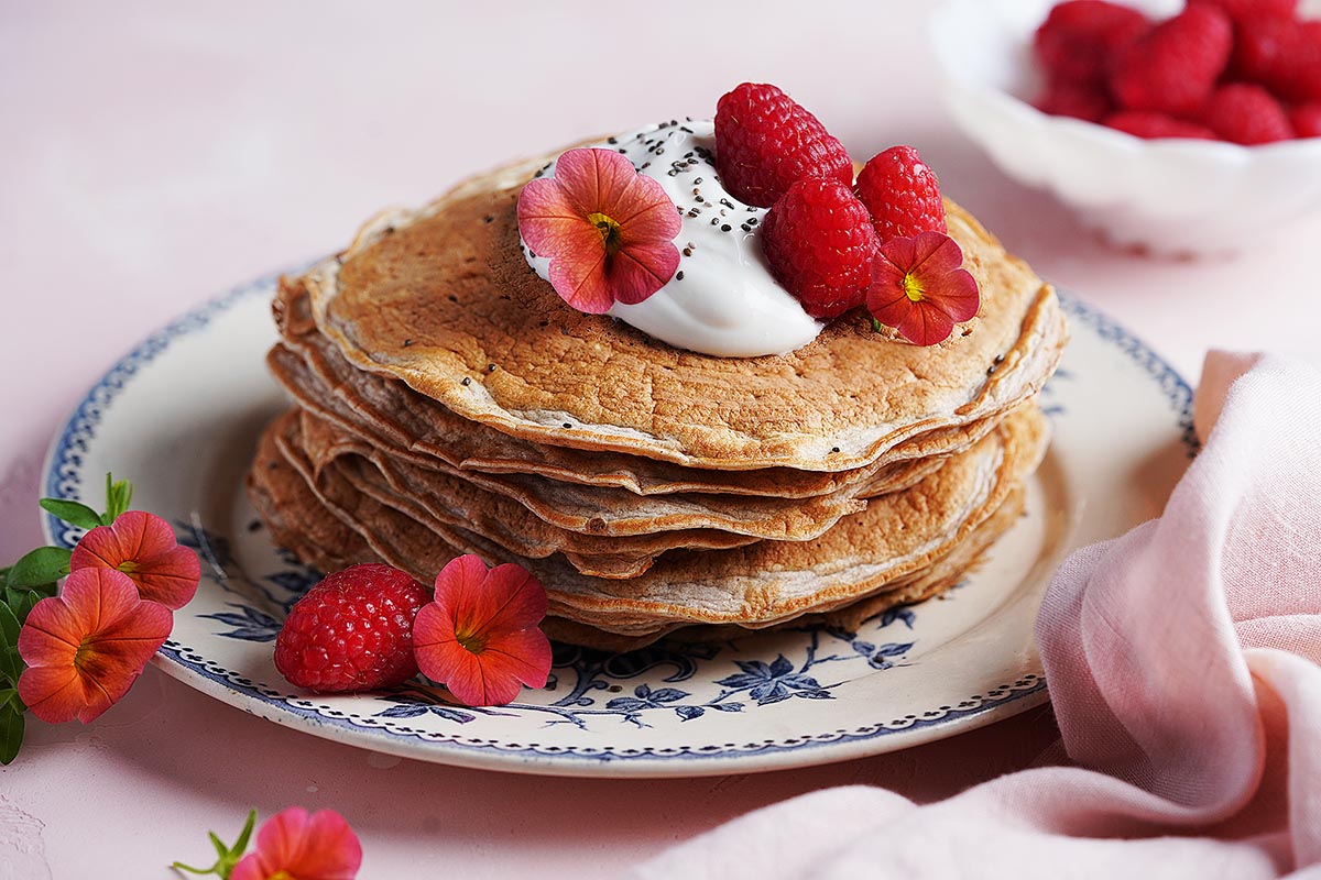A stack of Protein Pancakes Without Protein Powder topped with whipped cream, raspberries, chia seeds, and edible flowers sits on a decorative plate. A pink napkin and a bowl of raspberries are nearby on a light pink background.
