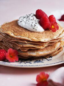 A stack of pancakes topped with whipped cream, chia seeds, raspberries, and edible orange flowers on a decorative plate.