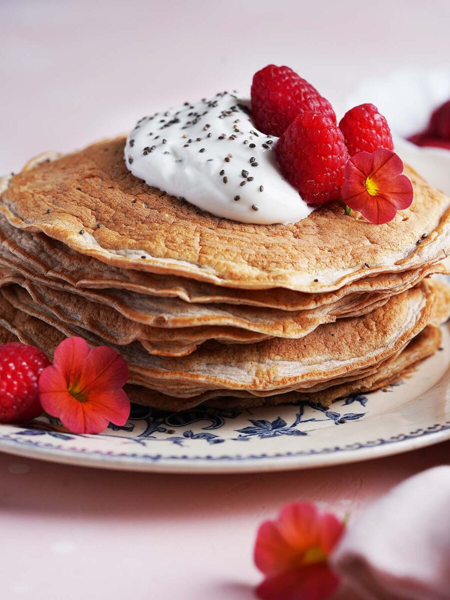 A stack of Protein Pancakes Without Protein Powder topped with whipped cream, chia seeds, raspberries, and edible orange flowers on a decorative plate.