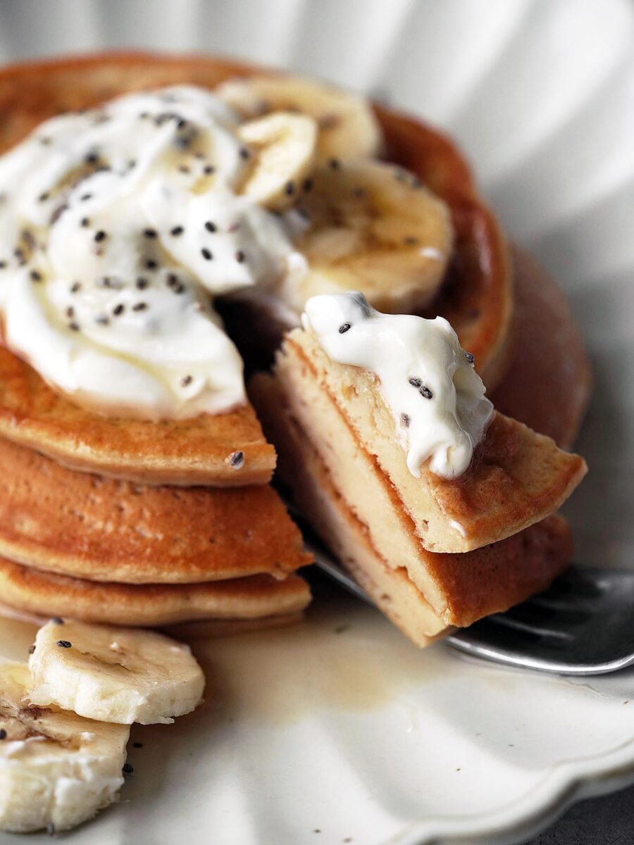 A stack of pancakes topped with sliced bananas, whipped cream, and chia seeds on a white plate. A fork holds a bite of pancake with bananas and cream.