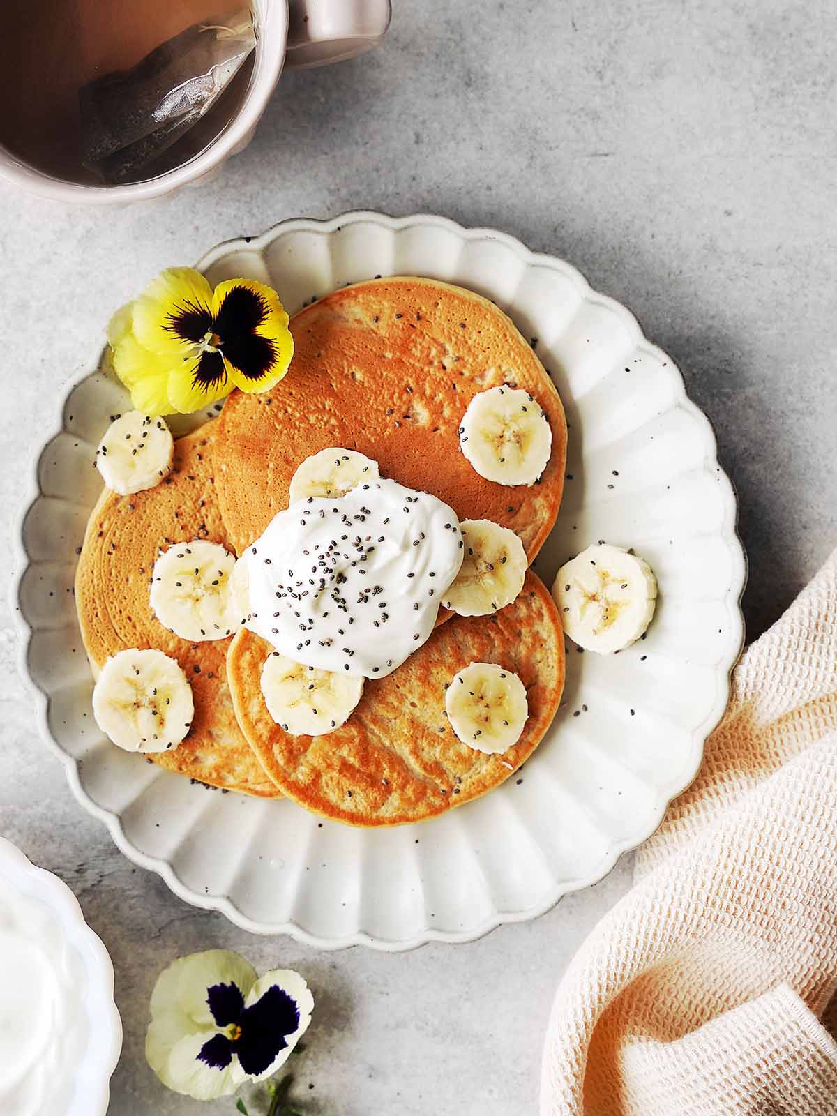 A scalloped white plate with three pancakes, banana slices, whipped cream, chia seeds, and edible flowers. A mug with tea and a beige cloth are beside the plate. The background is a light gray surface.