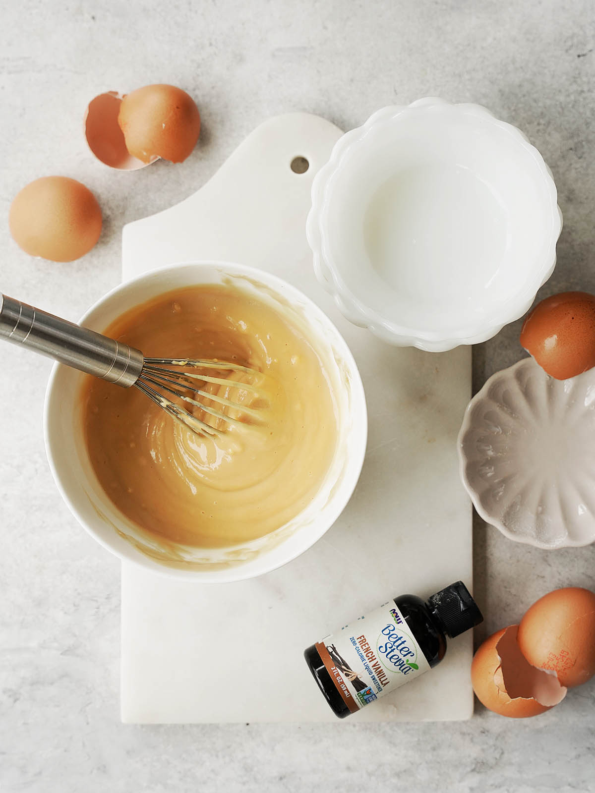 A bowl of batter with a whisk sits on a white board. Broken eggshells, an empty bowl, a small plate, and a bottle of vanilla extract surround it on a light countertop.