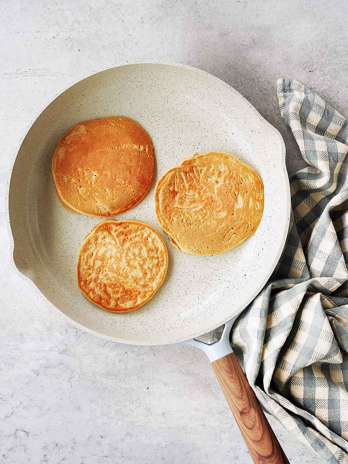 Three golden brown pancakes cooking in a white frying pan with a wooden handle. A gray and white checkered kitchen towel is placed next to the pan on a light-colored countertop.