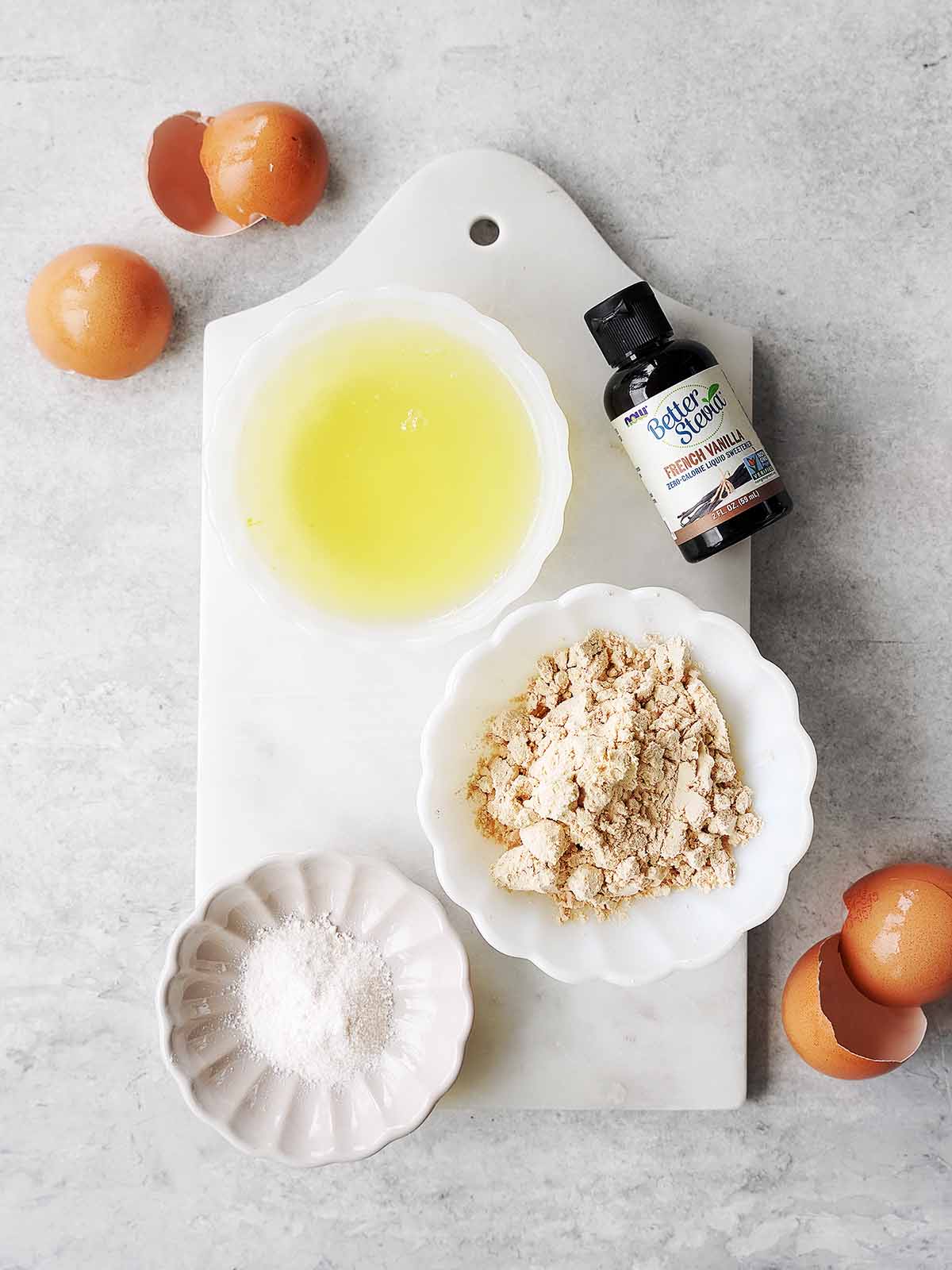 Four baking ingredients: egg whites in a bowl, almond flour in a dish, a small dish of sugar, and a bottle of vanilla extract, arranged on a white board, with empty eggshells around.
