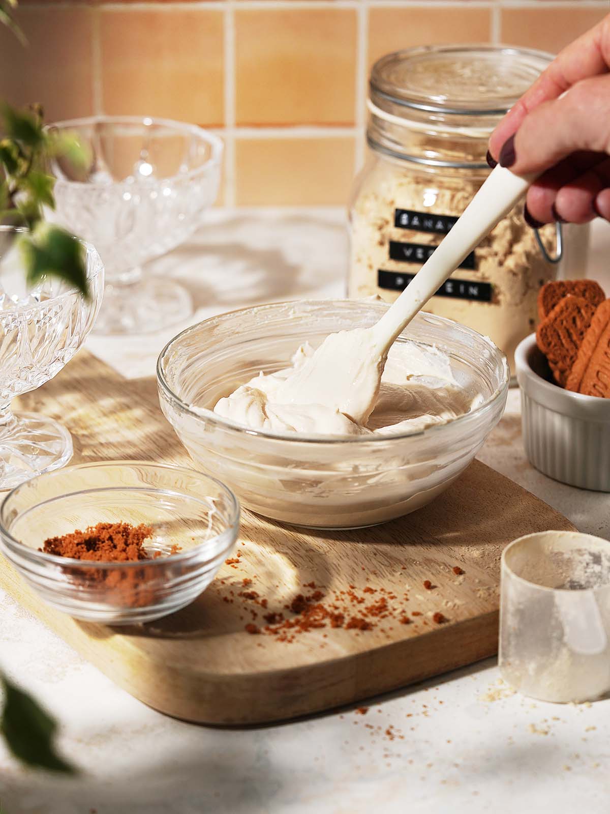 A hand uses a spatula to mix creamy dessert in a glass bowl on a wooden board. Nearby are crumb cookies, a jar of sugar, and small bowls with crushed cookies and powder.