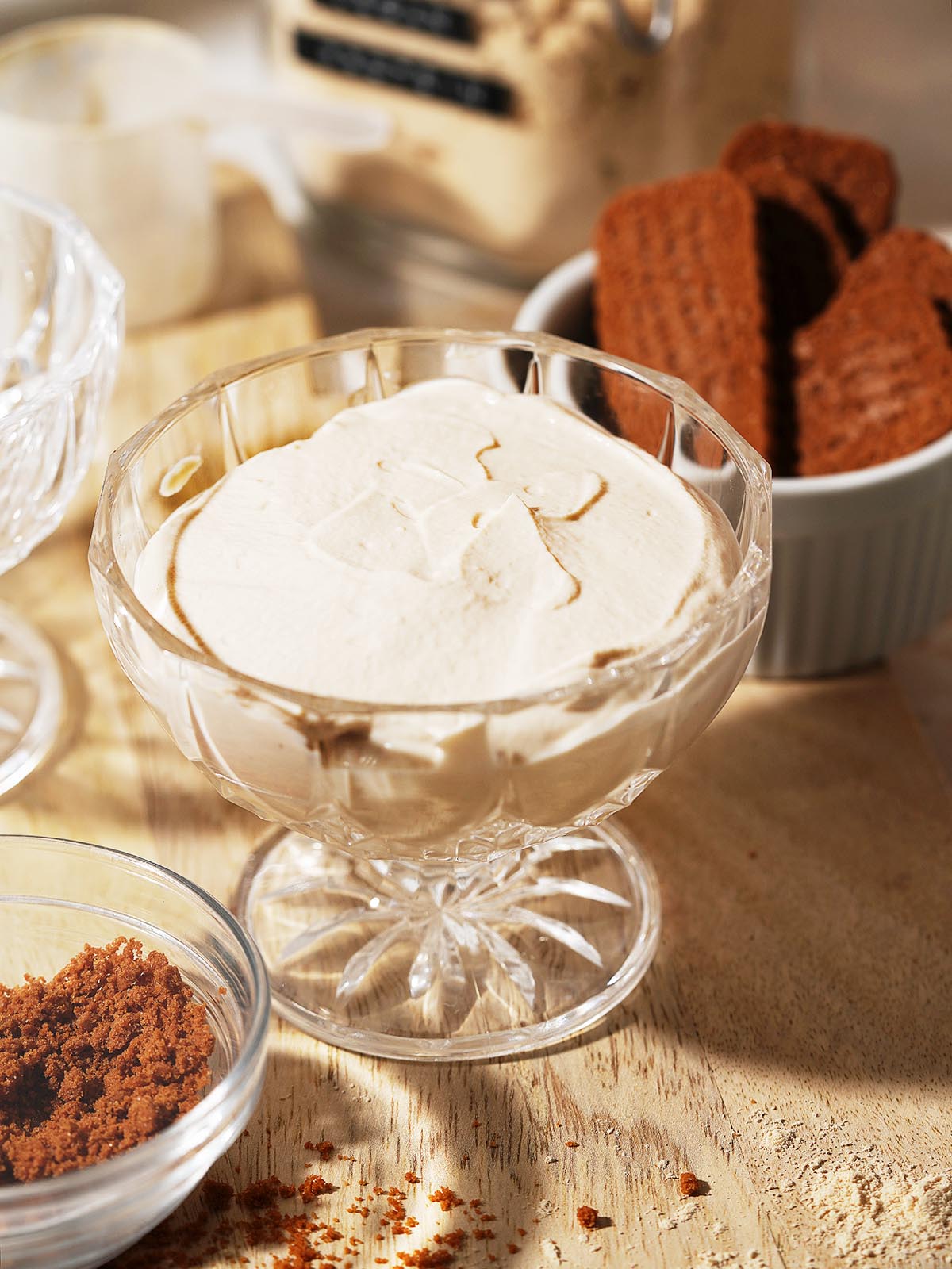 A glass dessert dish filled with creamy, light brown Protein Cheesecake sits on a wooden surface. Nearby are a bowl of chocolate cookies and a small bowl of crumbled chocolate, with soft sunlight illuminating the scene.